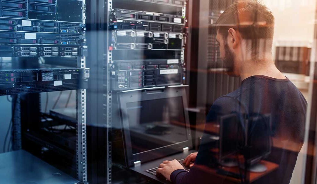 A person uses a laptop while standing in a server room, emphasizing the importance of in-house IT staff. Several server racks and networking equipment are visible in the background, showcasing the infrastructure crucial for business scalability.