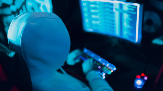 In a dimly lit room, a person in a hoodie works at a computer with code on the screen, focusing intently on addressing vulnerabilities and enhancing data protection.